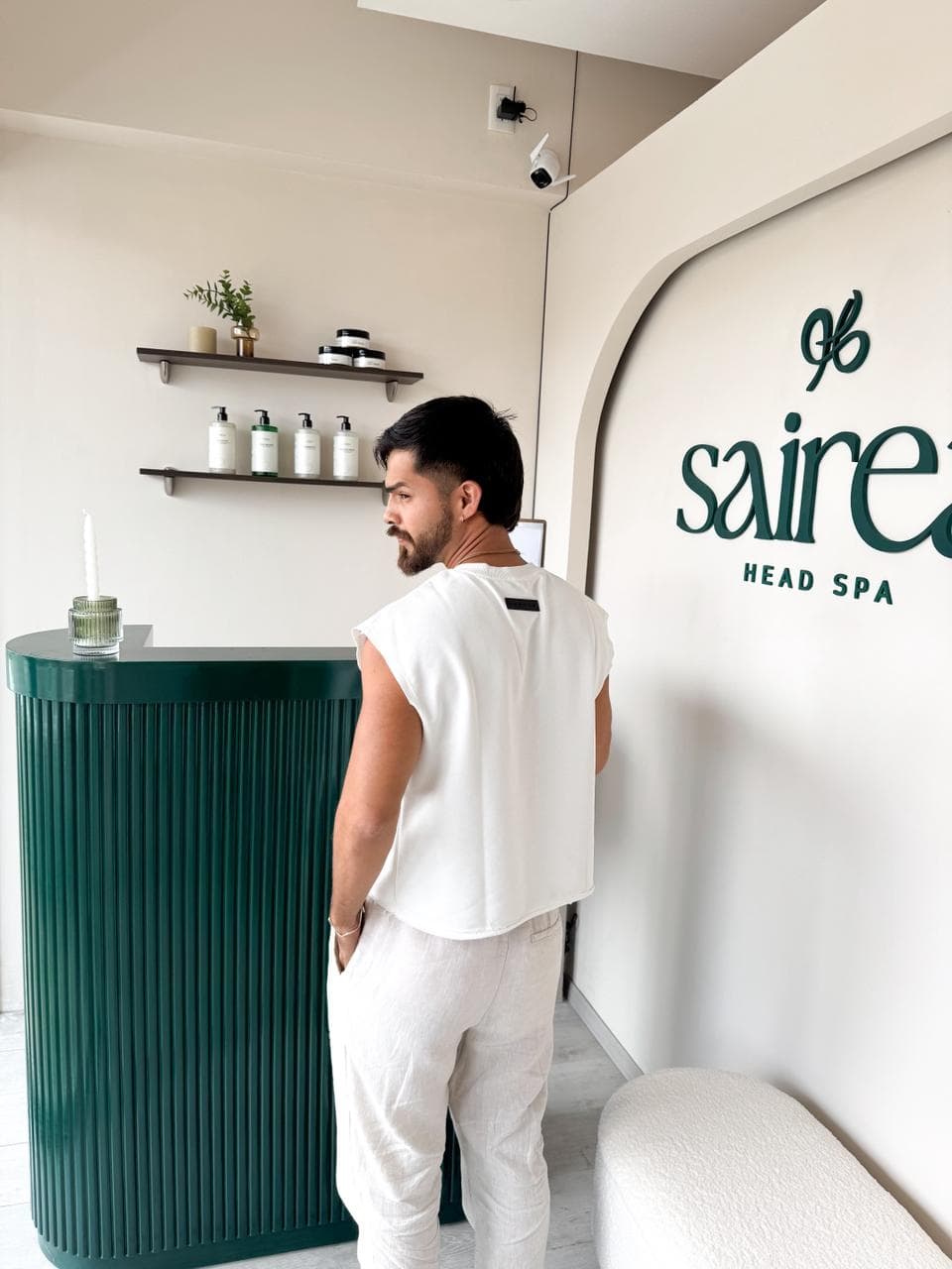 Recepción de Sairea Head Spa con su logo verde sobre pared blanca y un banco de bouclé